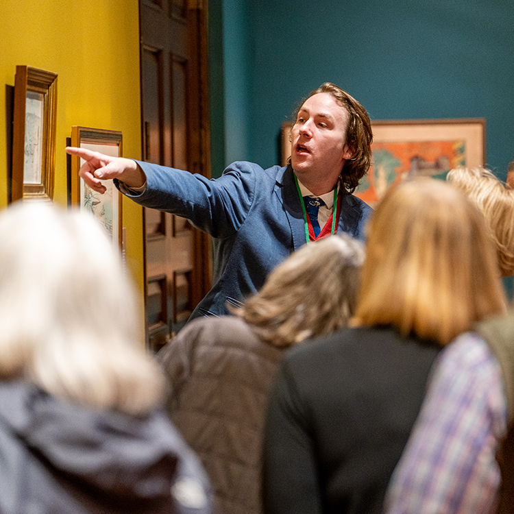 A guide points to a painting in an art museum as a crowd listens.