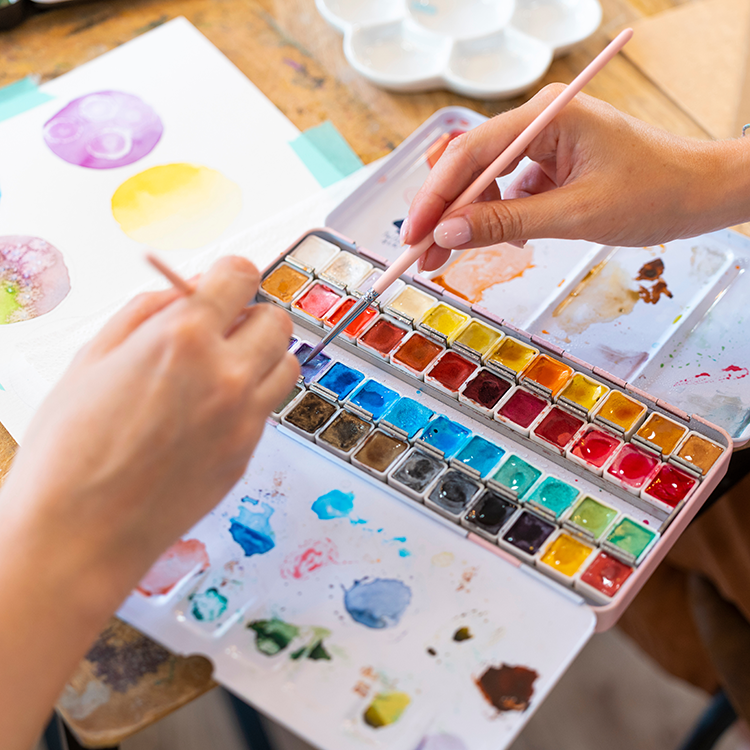 Two hands dip paintbrushes into a watercolor palette