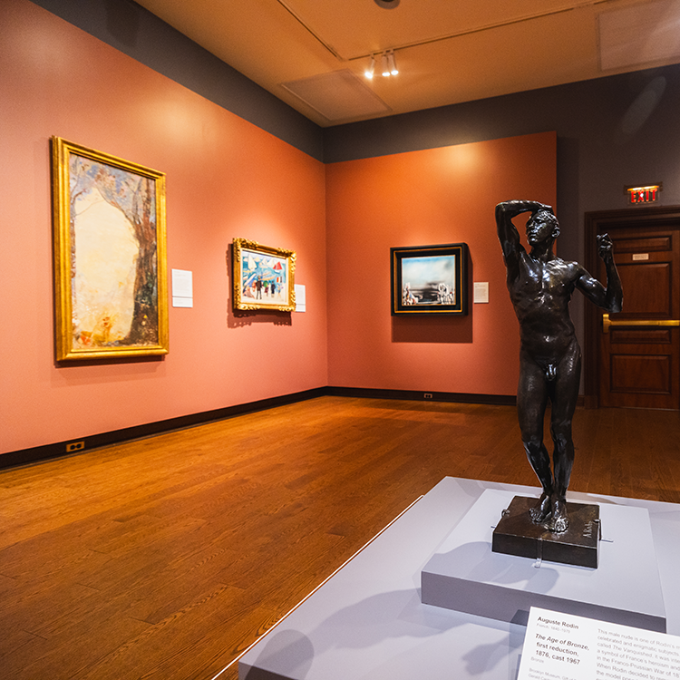 A sculpture of a man stands in an art museum. Paintings hang on the walls in the background.
