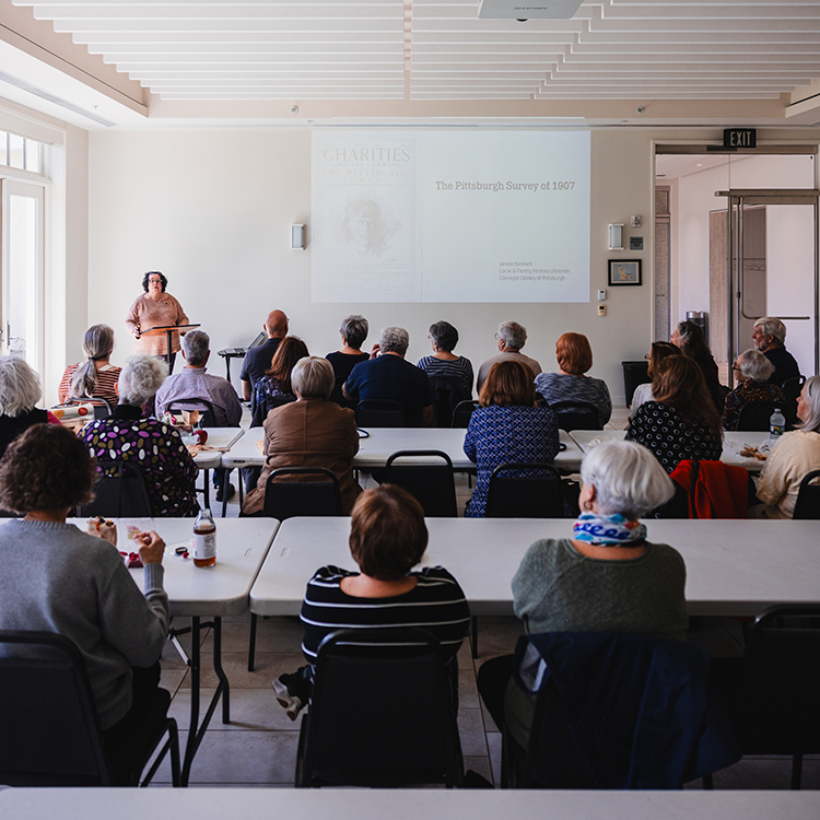 An audience of people sit at tables and listen to a lecturer give a presentation.